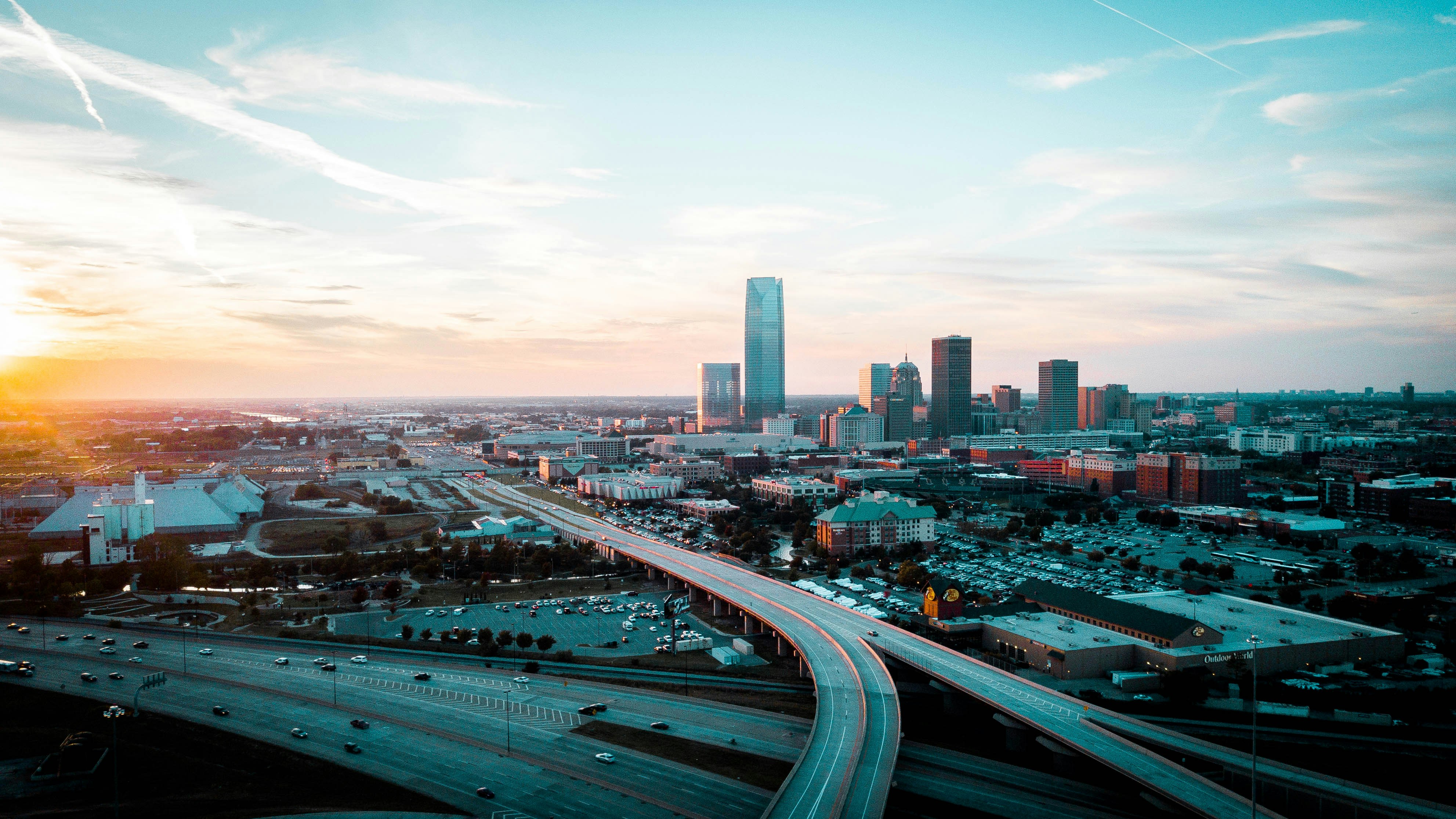 The Oklahoma City skyline at sunset, with the Devon Energy Center tower rising over downtown and a highway curving in toward the city.