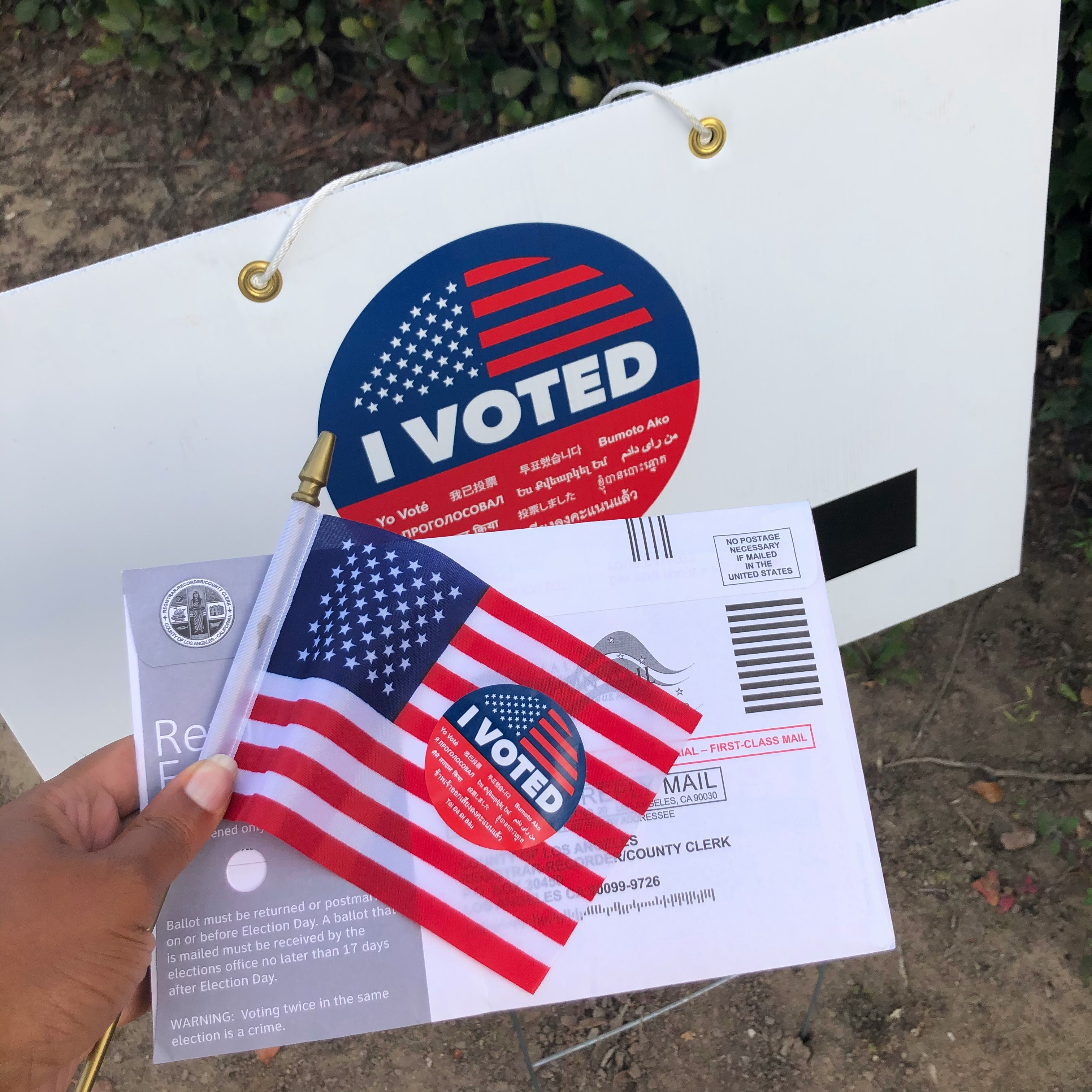 A voter holding a small American flag, a vote-by-mail ballot envelope, and an 'I Voted' sticker outside a polling place.
