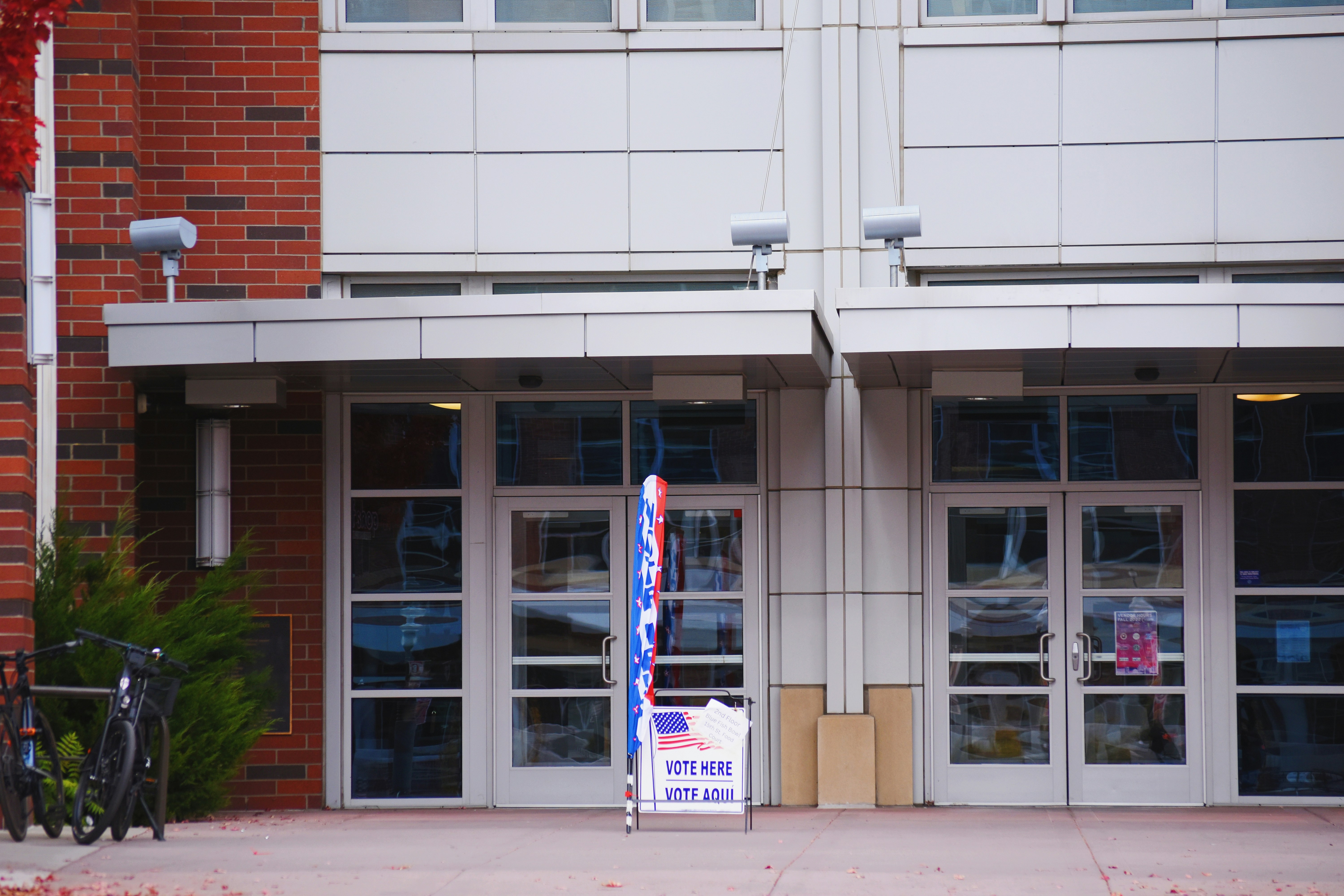 A quiet polling place entrance with a 'Vote Here / Vote Aquí' sign and red, white, and blue bunting at the door.